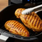 Gros plan sur un Airfryer ouvert avec deux blancs de poulet en train de cuire, dorés et légèrement croustillants, avec une pince soulevant un morceau pour révéler l’intérieur juteux.
